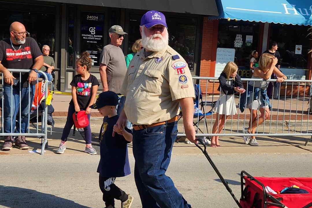 Photo of a male-presenting scout youth and male-presenting scout adult holding hands and pulling a cart, walking on a main street. Youth is wearing a lions cub tee and adult is wearing a scouting uniform shirt with several badges and a RSR baseball cap. There are bystanders in the background watching the procession.