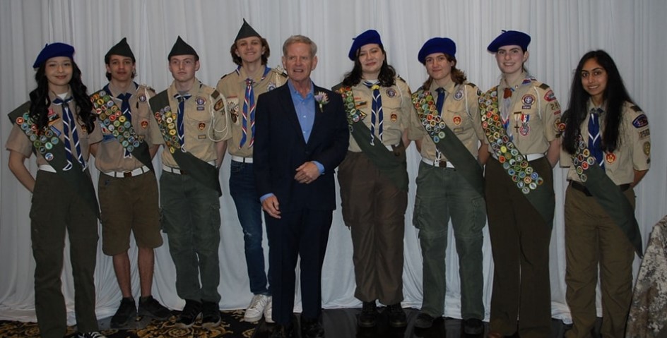 Group photo of female and male=presenting scouts standing next to an elderly male-presenting gentlemen in a suit. The scouts are wearing their uniforms, with full regalia, including badges and hats.