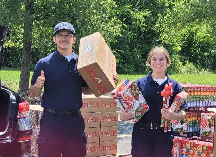 Photo of two youth holding snack boxes and cereal boxes. There is a pallet of food behind them and they are smiling for the camera.
